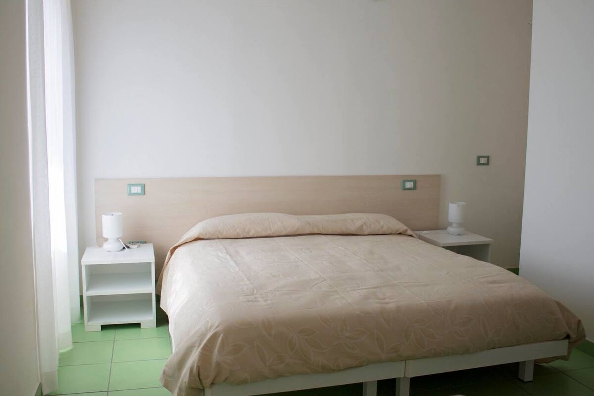 Un lit bien fait, recouvert d’une couvre-lit beige, avec deux tables de chevet sur lesquelles sont posées des lampes.