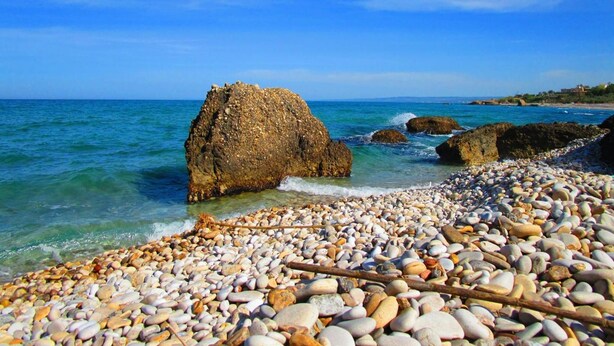 Una spiaggia di ciottoli con acque cristalline e un promontorio roccioso.