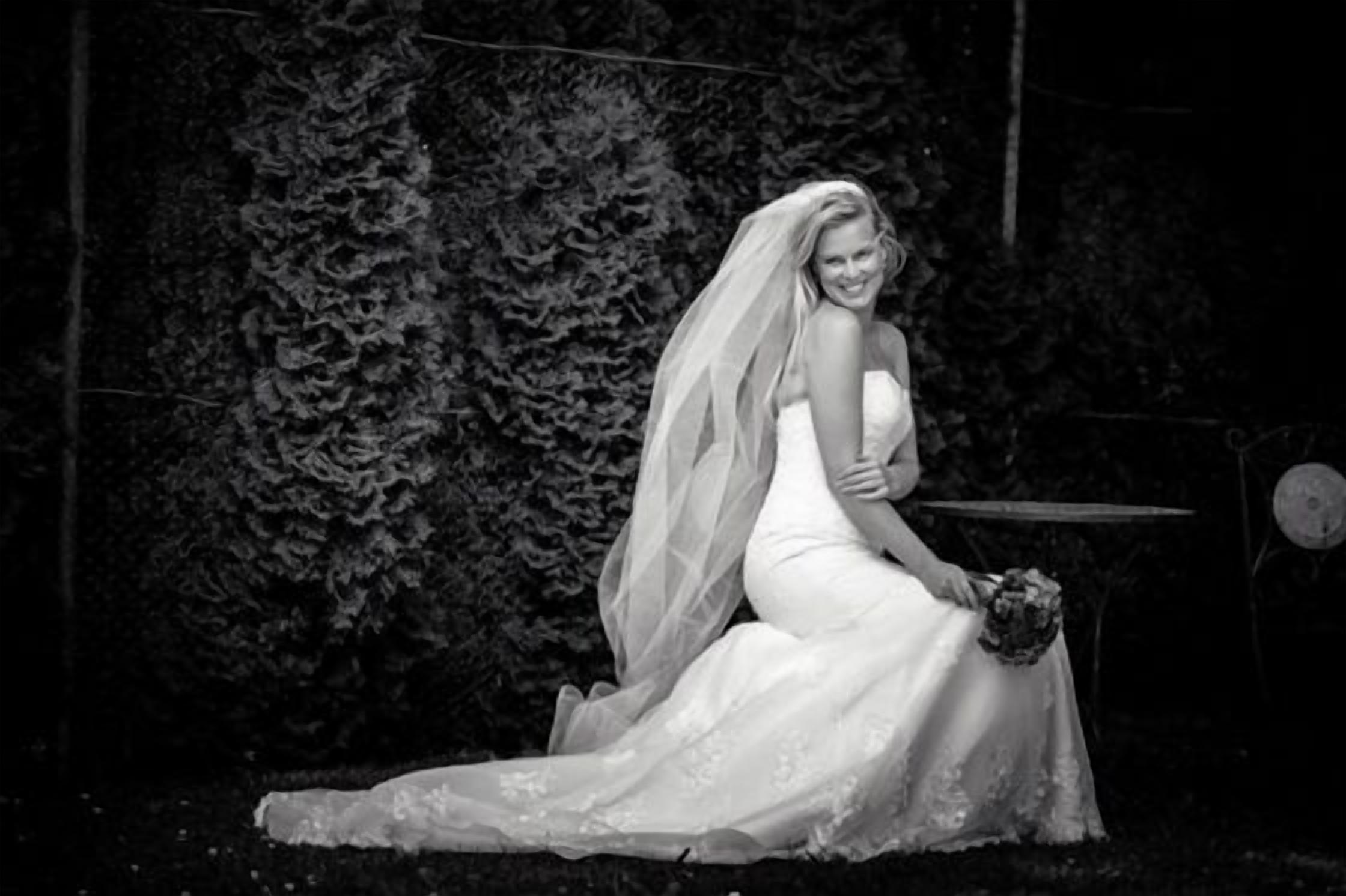 Eine Hochzeitsszene mit einer Braut im weißen Kleid, die einen Blumenstrauß hält und vor dichtem Grünwerk sitzt.