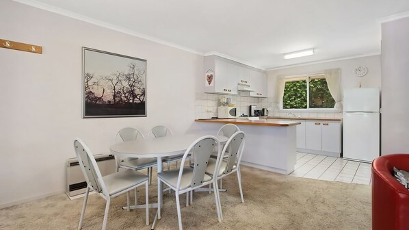 Uma cozinha compacta com armários brancos, uma mesa de jantar com cadeiras, uma geladeira e uma janela com vista para o verde.