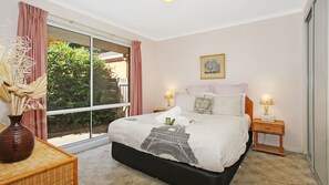 Um quarto com uma cama, duas mesas de cabeceira, uma janela com cortinas, um vaso com plantas secas e um quadro na parede.