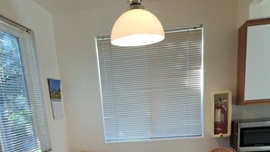 Ein Essbereich mit einem runden Holztisch, vier Holzstühlen, einer Deckenleuchte, einem Fenster mit Jalousien und einer Mikrowelle in einem Schrank.
