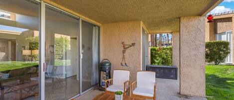 A covered patio with a glass door leading to a living area, a wooden table, two white chairs, and a small plant.