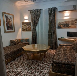 A living room with patterned sofas, a glass coffee table, wall-mounted artwork, and an air conditioning unit. - ALGER CENTRE (CHAMANEUF) APPARTMENT A LOUER AVEC INTERNET/DOUBLE VITRAGE (Alger)