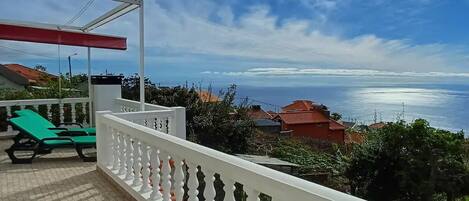 A balcony with a white balustrade, green lounge chairs, and a view of the sea and buildings in the distance.