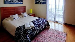 Slaapkamer met een houten bed, twee schilderijen aan de muur, een raam met gordijnen en een klein nachtkastje met een lamp.