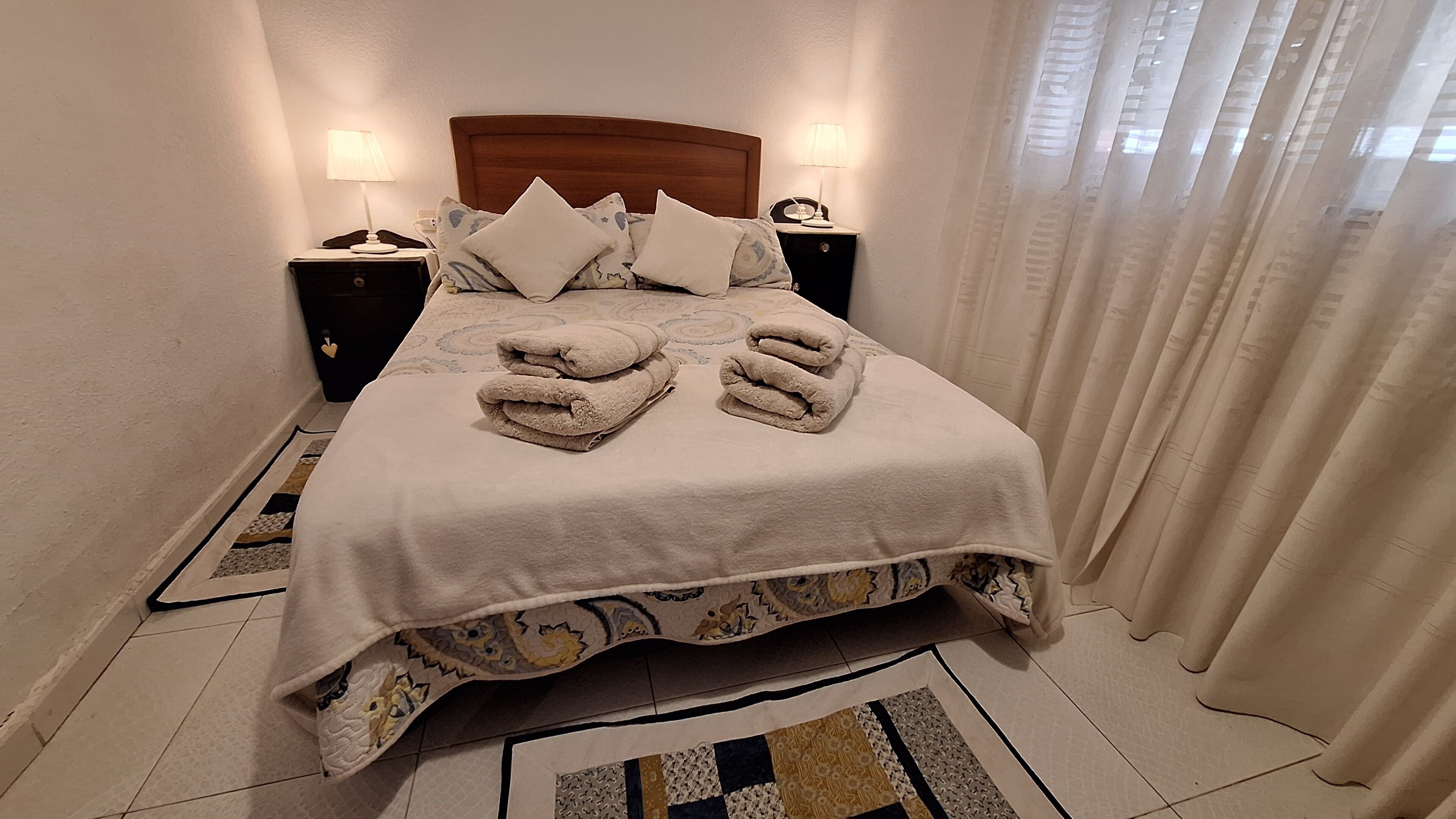 Een net opgemaakt bed met een bedspread met patroon en kussens, twee nachtkastjes met lampen, en een decoratief vloerkleed.