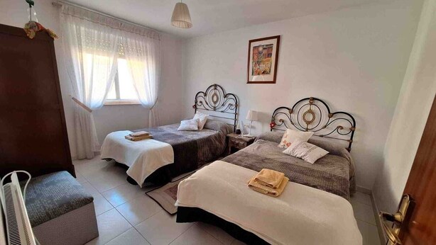 Un dormitorio con dos camas, una ventana con cortinas, una mesita de noche con lámpara y un cuadro en la pared.