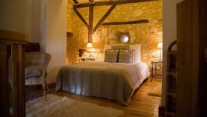 Un dormitorio acogedor con pared de piedra, vigas de madera, cama con almohadas estampadas con corazones, una silla de mimbre y una mesita de noche con una lámpara.