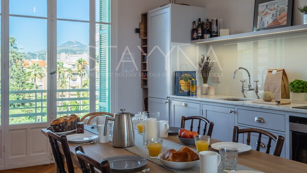 A kitchen with a dining table set for breakfast, a view of the outdoors, and a cabinet with kitchen items.