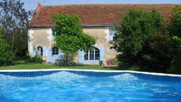 Una piscina con borde de azulejos, un edificio de techo rojo y paredes blancas, y árboles verdes al fondo.