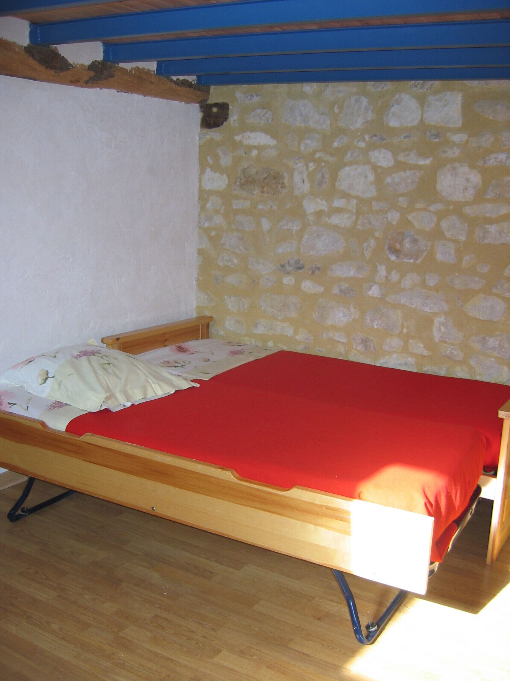 Una cama individual con una colcha roja, estructura de madera y un cojín estampado con motivos florales. La habitación tiene una pared de piedra y suelo de madera.