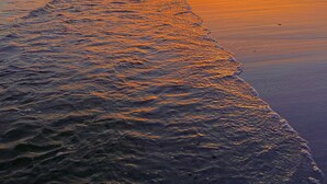 Auringonlasku tyynen joen yllä, näkyvissä veneen jättämä aallokko, kaukainen rantaviiva ja rakennuksia.