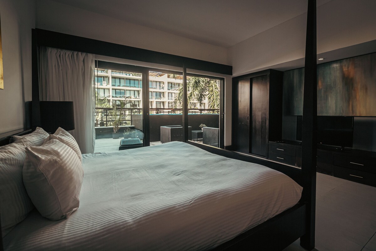 A modern bedroom with a large bed, a TV, and a balcony with a view of buildings and palm trees.