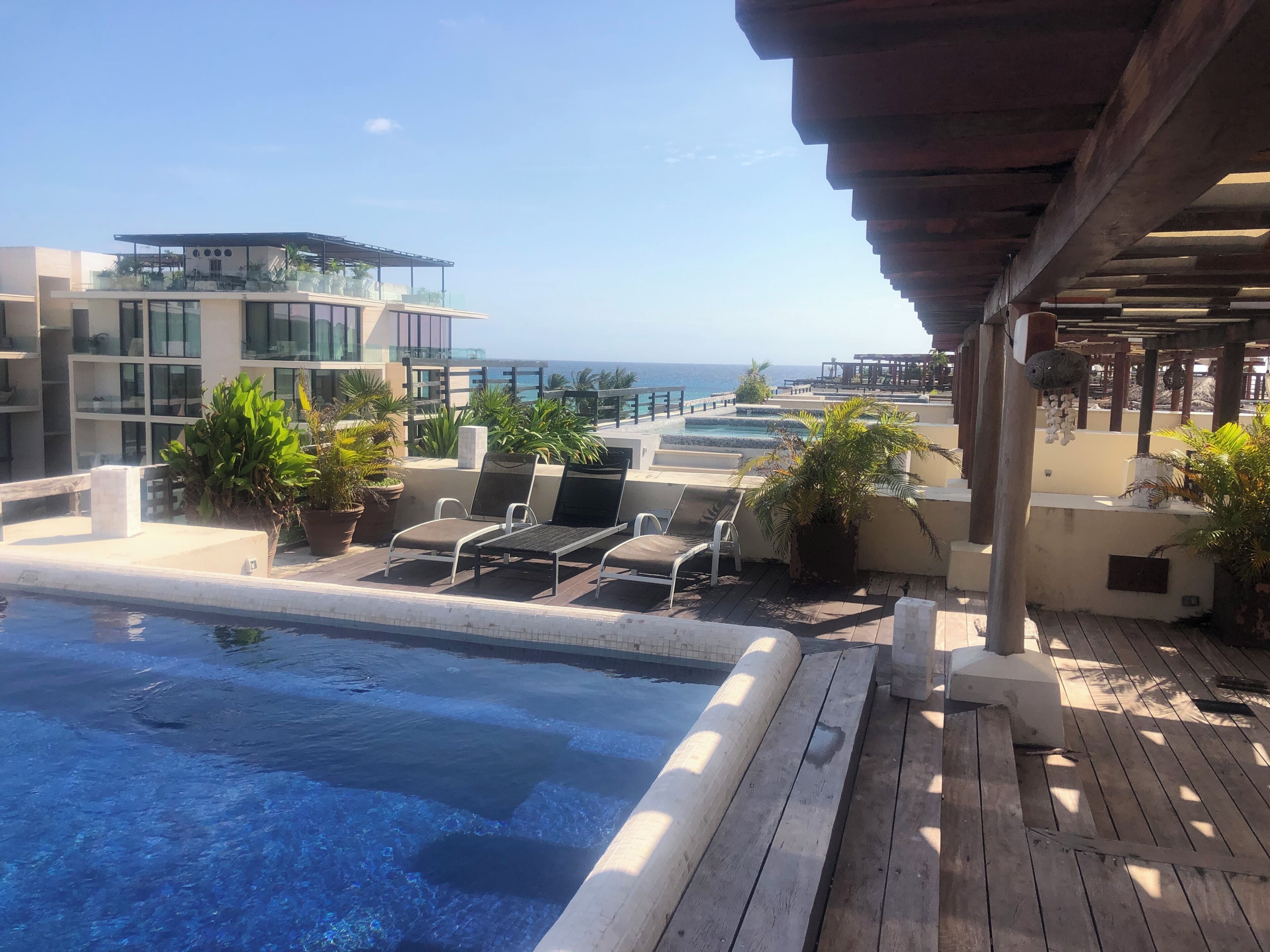A rooftop pool area with lounge chairs and potted plants, overlooking a body of water and a building with large windows.