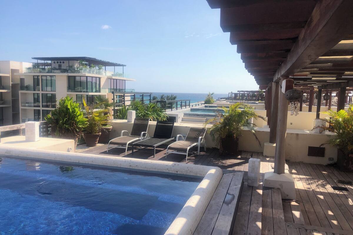 A rooftop pool area with lounge chairs and potted plants, overlooking a body of water and a building with large windows.