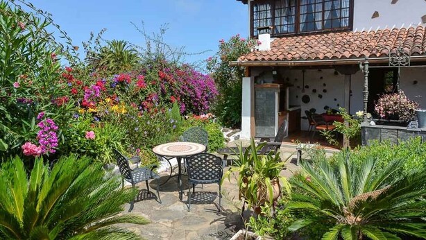 Una casa con techo de tejas, un patio con mesa y sillas redondas, y un jardín con diversas plantas y flores.