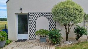 Une maison dotée d’une grande fenêtre, d’une porte ornée d’un motif en treillis noir et d’un petit jardin où poussent diverses plantes et fleurs.