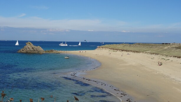 Une plage avec une eau d’un bleu limpide, un rivage sablonneux et des bateaux à l’horizon.
