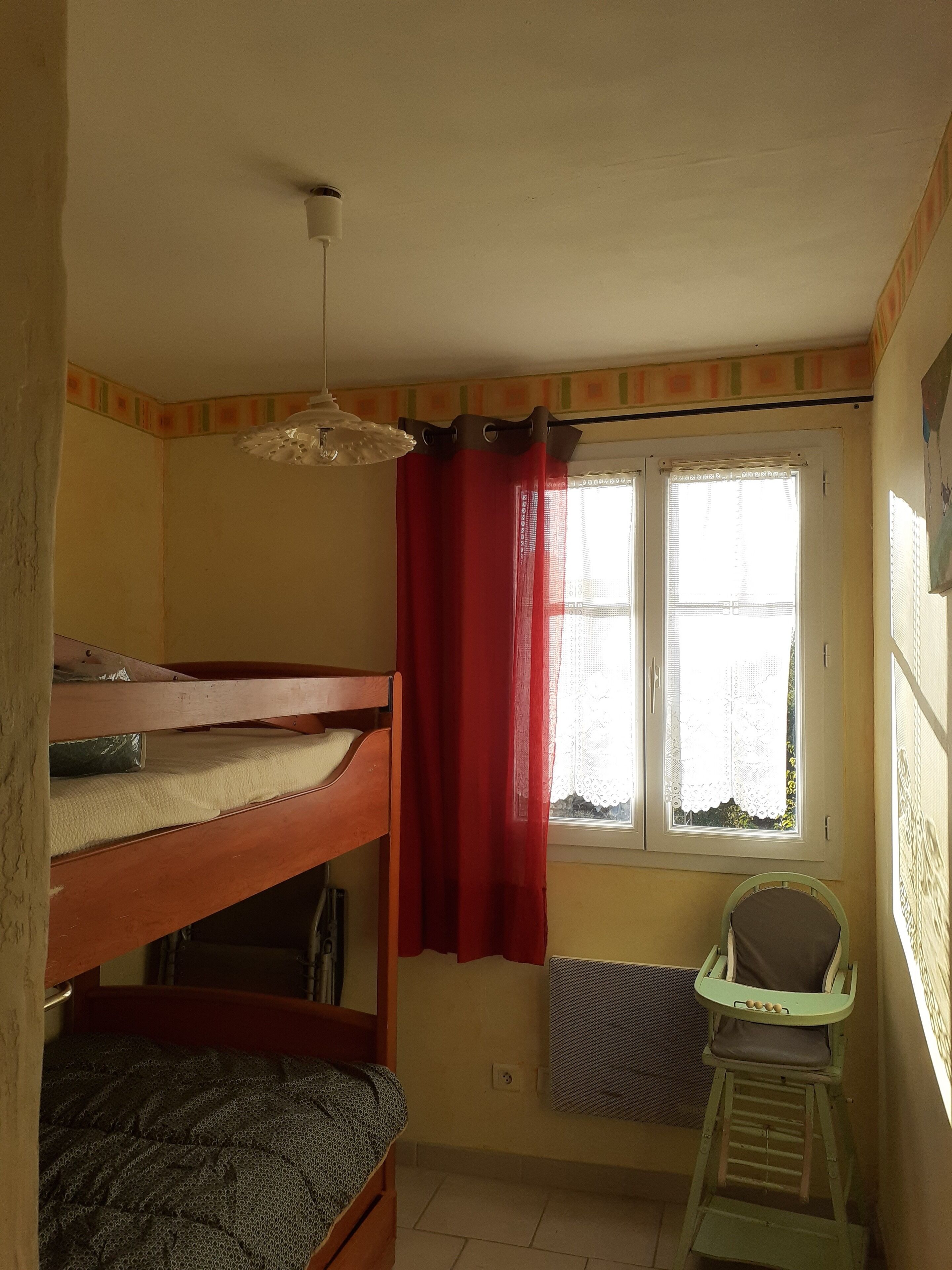 A dormitory room with bunk beds, a window with red curtains, a radiator, and a small chair.