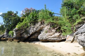 Une plage rocheuse aux eaux cristallines, un escalier menant au sommet d’une falaise et une végétation luxuriante.