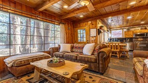 A cozy cabin interior with a wooden ceiling, large windows, a sofa set, a dining table, and a kitchen area. - Family cabin: Hot tub, firepit, game room, fenced, BBQ, movie lineup (Big Bear)