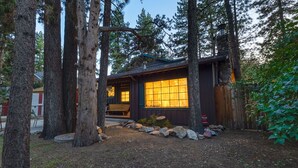 A rustic cabin with a large window, surrounded by tall trees and a stone fireplace. - Family cabin: Hot tub, firepit, game room, fenced, BBQ, movie lineup (Big Bear)