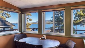 Quarto com uma mesa redonda e duas cadeiras, grandes janelas com vista para um lago e árvores, e teto de madeira.