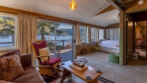 Interior acolhedor de uma cabana com piso de madeira, um sofá, uma poltrona, uma mesa pequena com um livro e uma cama. Grandes janelas oferecem vista para um lago e árvores.