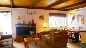 Un salon chaleureux avec un plafond en bois, une télévision, un canapé, une table basse et un coin repas comprenant une table et des chaises.