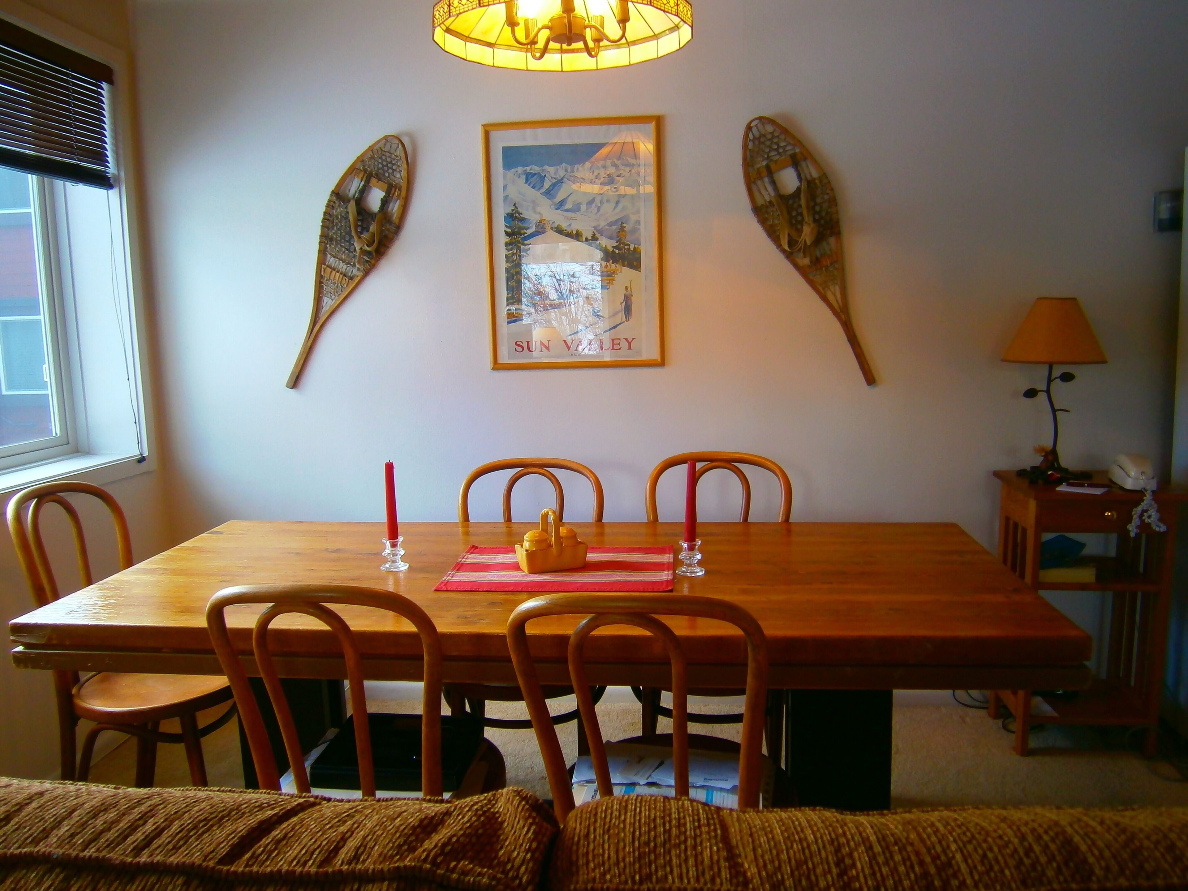 Uma área de jantar com mesa de madeira, quatro cadeiras, um quadro emoldurado e uma luminária em uma mesinha lateral.