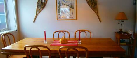 Uma área de jantar com mesa de madeira, quatro cadeiras, um quadro emoldurado e uma luminária em uma mesinha lateral.