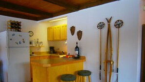 Une cuisine avec des armoires en bois, un réfrigérateur, un four à micro-ondes et une bouteille de vin posée sur le comptoir. On y trouve deux tabourets et des bâtons de ski appuyés contre le mur.