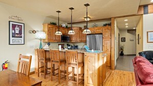 Una cocina con armarios de madera, una isla central con taburetes, un microondas y un refrigerador. Hay una mesa de comedor con sillas y un extintor en la pared.