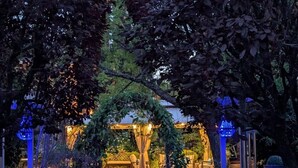 Un camí de jardí exterior que condueix a una zona d'seure coberta amb taules i cadires, envoltada d'arbres i il·luminada amb llums blaves.