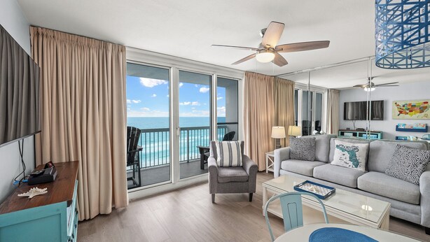 A modern living room with a sofa, armchair, and coffee table, large windows with curtains, and a ceiling fan.