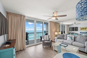 A modern living room with a sofa, armchair, and coffee table, large windows with curtains, and a ceiling fan. - CONDO IS ON THE BEACH! 
 240+ 5-star reviews - Tiki Bar - 3 Pools - 2 Hot Tubs (Destin)