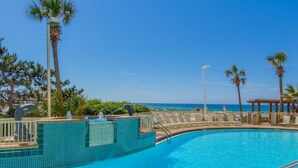 A clear blue pool with a tiled edge, surrounded by lounge chairs and palm trees, with a view of the ocean.
