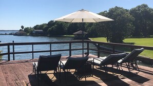 A wooden deck with outdoor furniture and a large umbrella, overlooking a body of water and a grassy area.