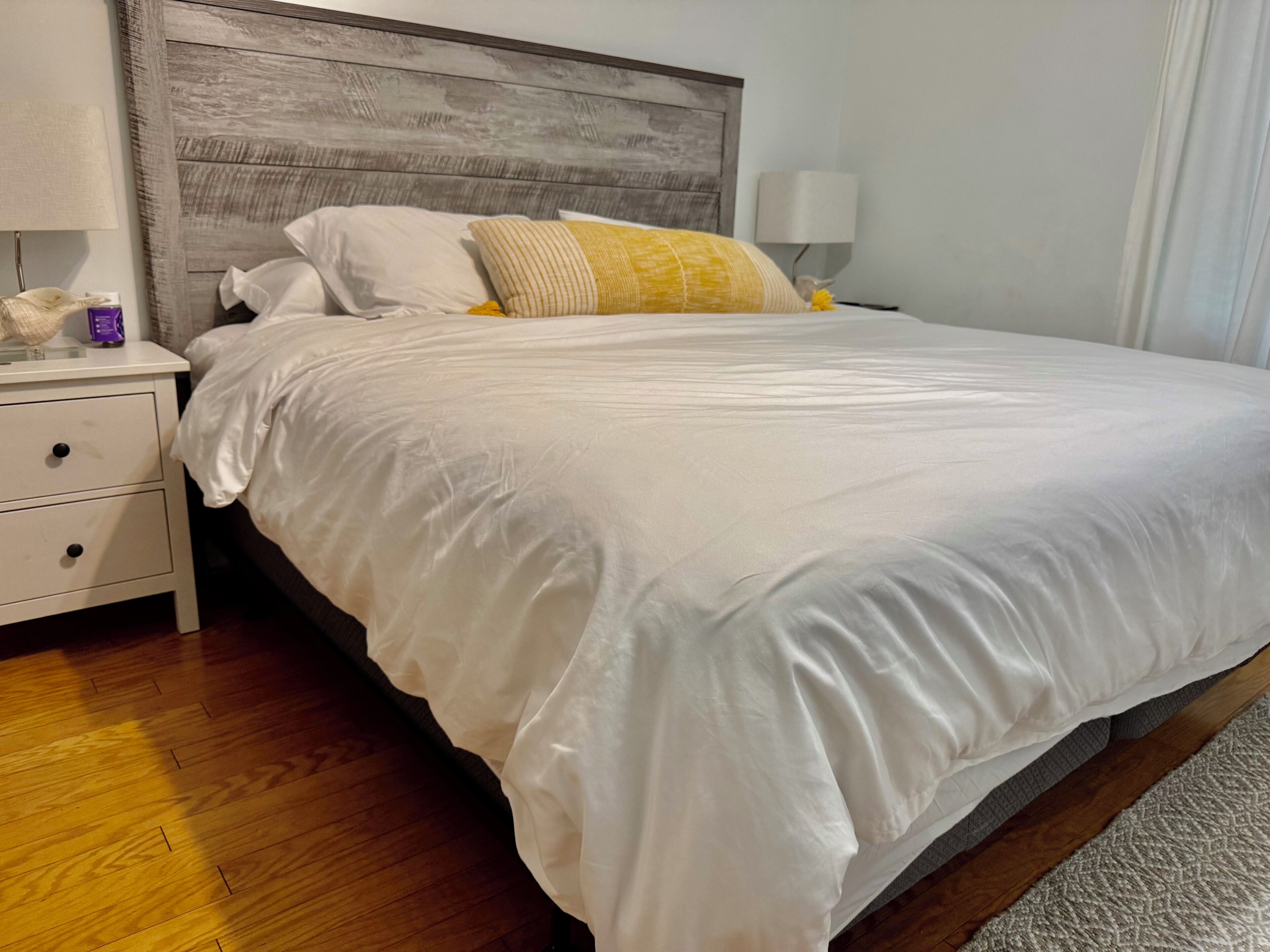A neatly made bed with white bedding and a wooden headboard, a bedside table with a lamp, and a wooden floor.