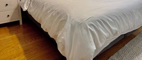 A neatly made bed with white bedding and a wooden headboard, a bedside table with a lamp, and a wooden floor.