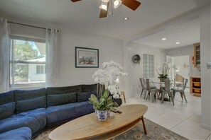 Ein Wohnzimmer mit einem blauen Sofa, einem Holztisch mit einer Vase voller weißer Blumen, einem Essbereich mit Tisch und Stühlen sowie einem Deckenventilator.