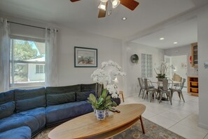 A living room with a blue sofa, a wooden coffee table with a vase of white flowers, a dining area with a table and chairs, and a ceiling fan.