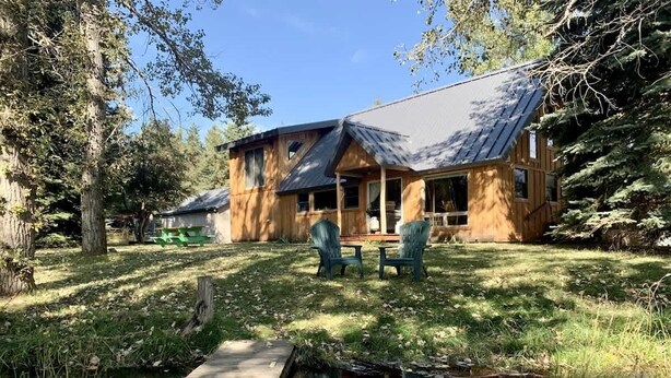 Une cabane en bois avec une véranda, un petit étang et deux chaises.
