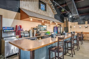 Uma área de bar com um balcão comprido, banquetas, uma máquina de Pepsi e uma cozinha ao fundo.