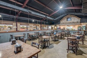 Um restaurante com várias mesas e cadeiras, um balcão com menu e uma estrutura de madeira rústica ao fundo.