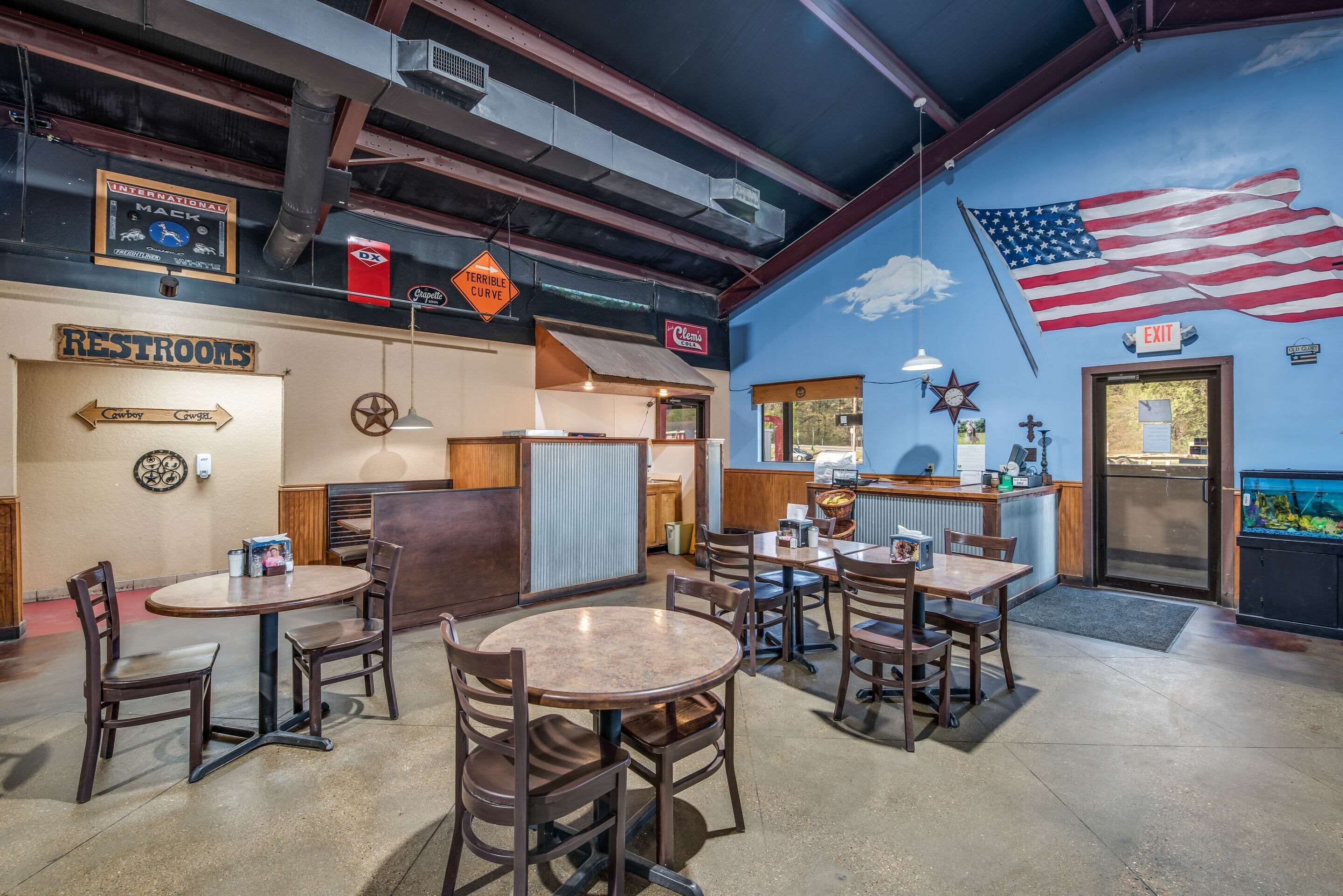 L’intérieur d’un restaurant, avec de nombreuses tables et chaises, une fresque représentant le drapeau américain, ainsi que des panneaux indiquant les toilettes et la sortie.