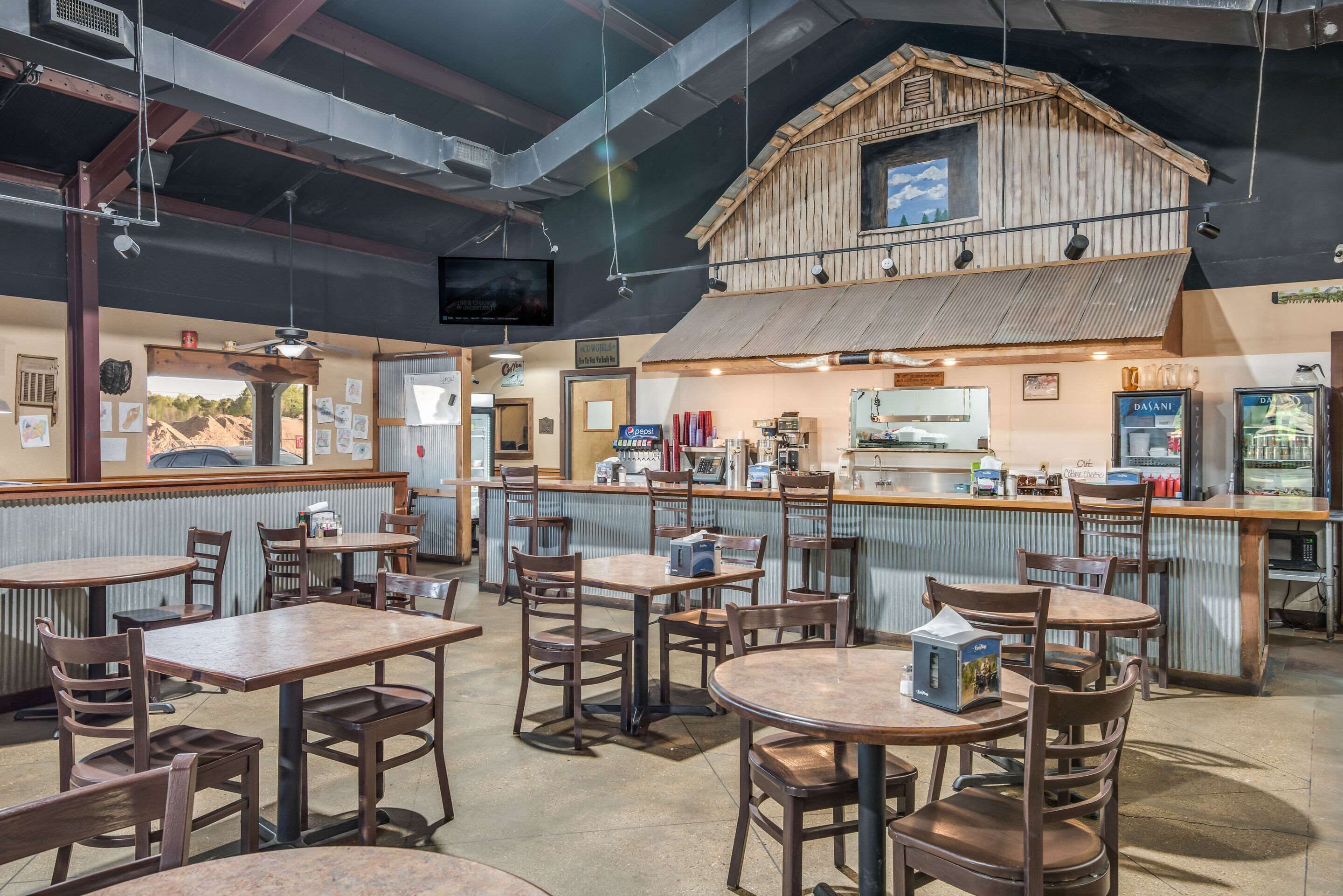 Interior de restaurante con mesas y sillas de madera, una barra con caja registradora y un televisor colgado en la pared.