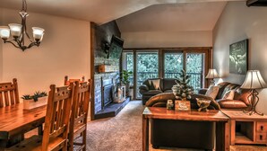 A cozy living room with a fireplace, a dining area with wooden furniture, and a balcony with a view of trees. - Breckenridge Peak 8 updated Ski in condo (Breckenridge)