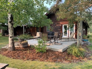 Relax in this cozy cabin with a patio area featuring a table, chairs, and fire pit, surrounded by a lawn and a tree.